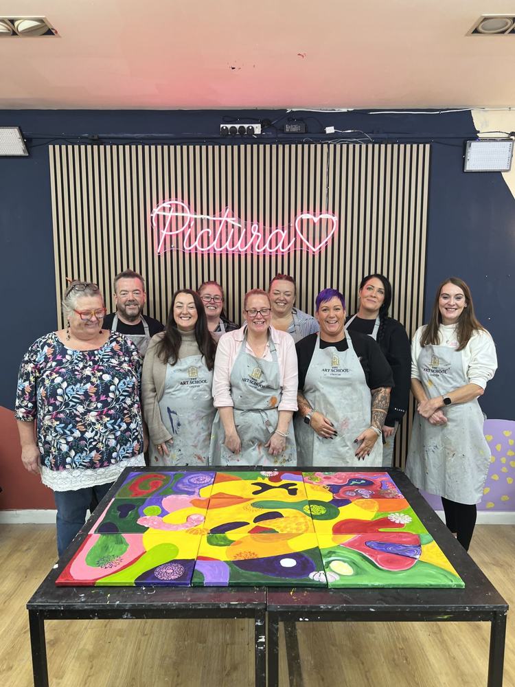 Nine colleagues in aprons standing with the giant nine-canvas painting they made together at their team day, under the Pictura neon sign