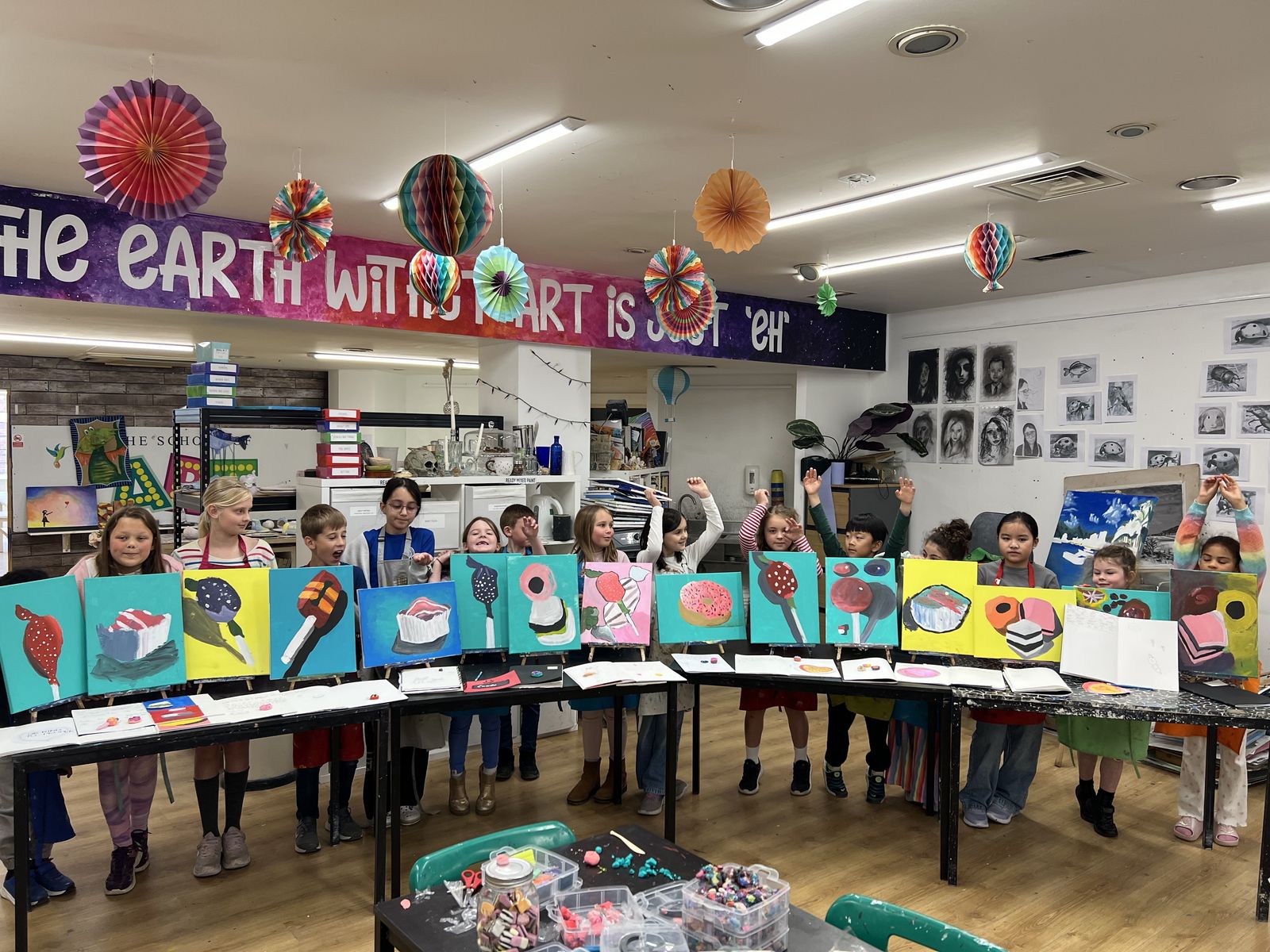 A full class of children in aprons cheering behind a long row of their finished colourful paintings in The Art School studio